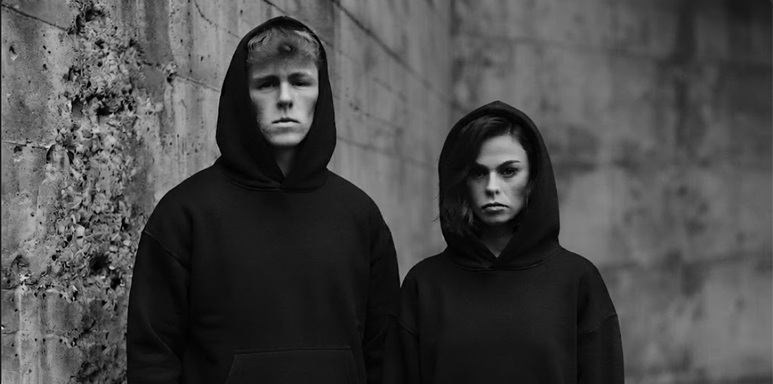 Two people in black Hagers hoodies against a concrete wall
